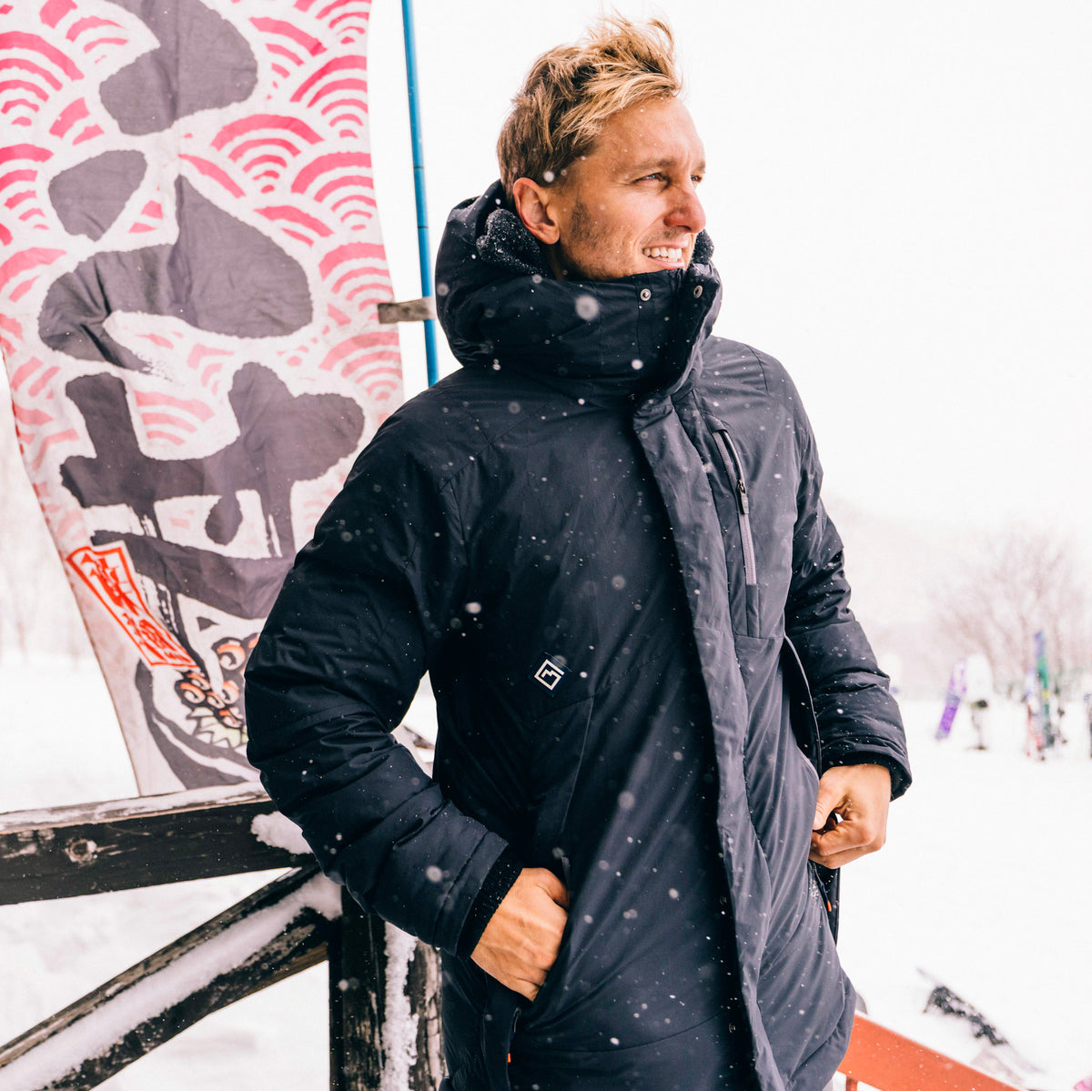 A man wearing a black hooded winter Cameron Pass Parka stands outdoors in the snow, leaning against a wooden post next to a decorative banner with red and gray wave patterns.