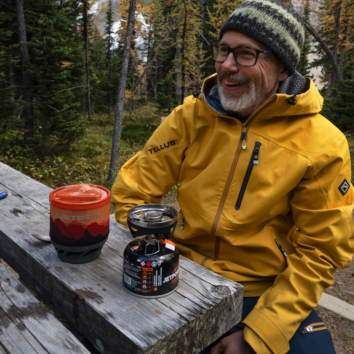 A man wearing a yellow TELLUS Long's Peak Waterproof Rain Shell jacket and a green knit beanie sits at a wooden picnic table outdoors in a forest, with a camping mug and a portable stove in front of him.