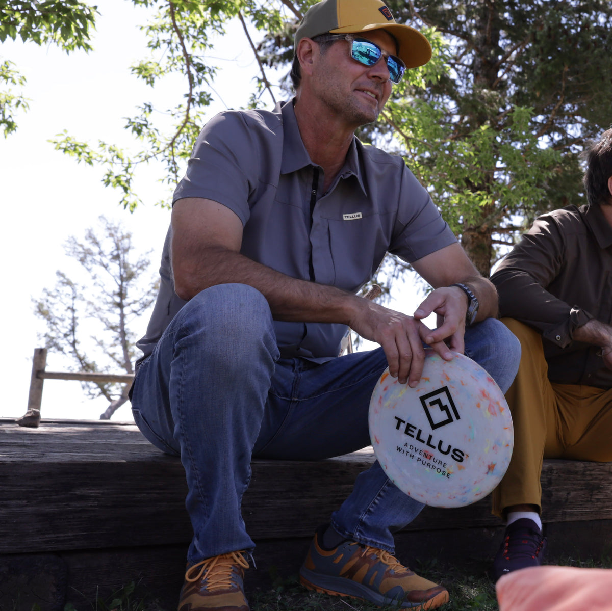Man sitting on a log wearing a gray Old Town Button Down shirt holding a Tellus frisbee,