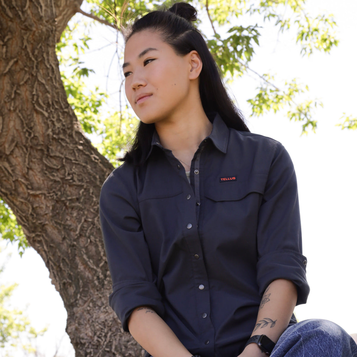 A woman with her hair in a bun sits outdoors next to a tree trunk, wearing a blue Foothills Flannel button-up shirt with a small orange Tellus logo on the chest pocket.