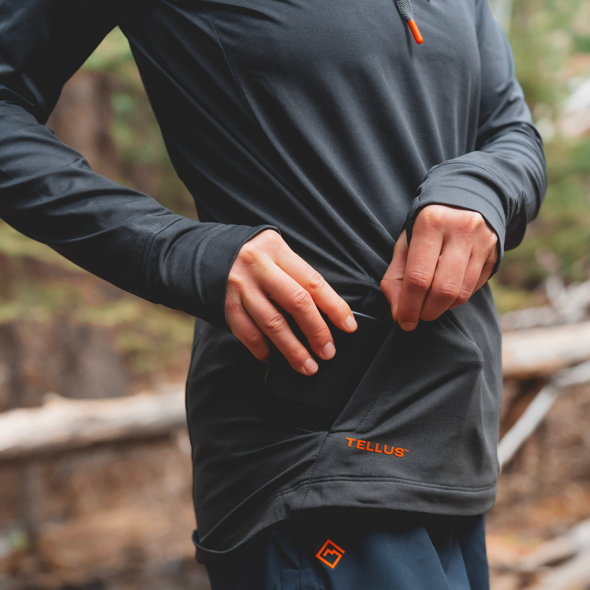 A close-up of a woman outdoors wearing a dark charcoal gray Horsetooth Performance Hoodie athletic top with an orange TELLUS logo near the hem, highlighting zip side pocket and thumbholes. The background is a blurred natural forest setting.