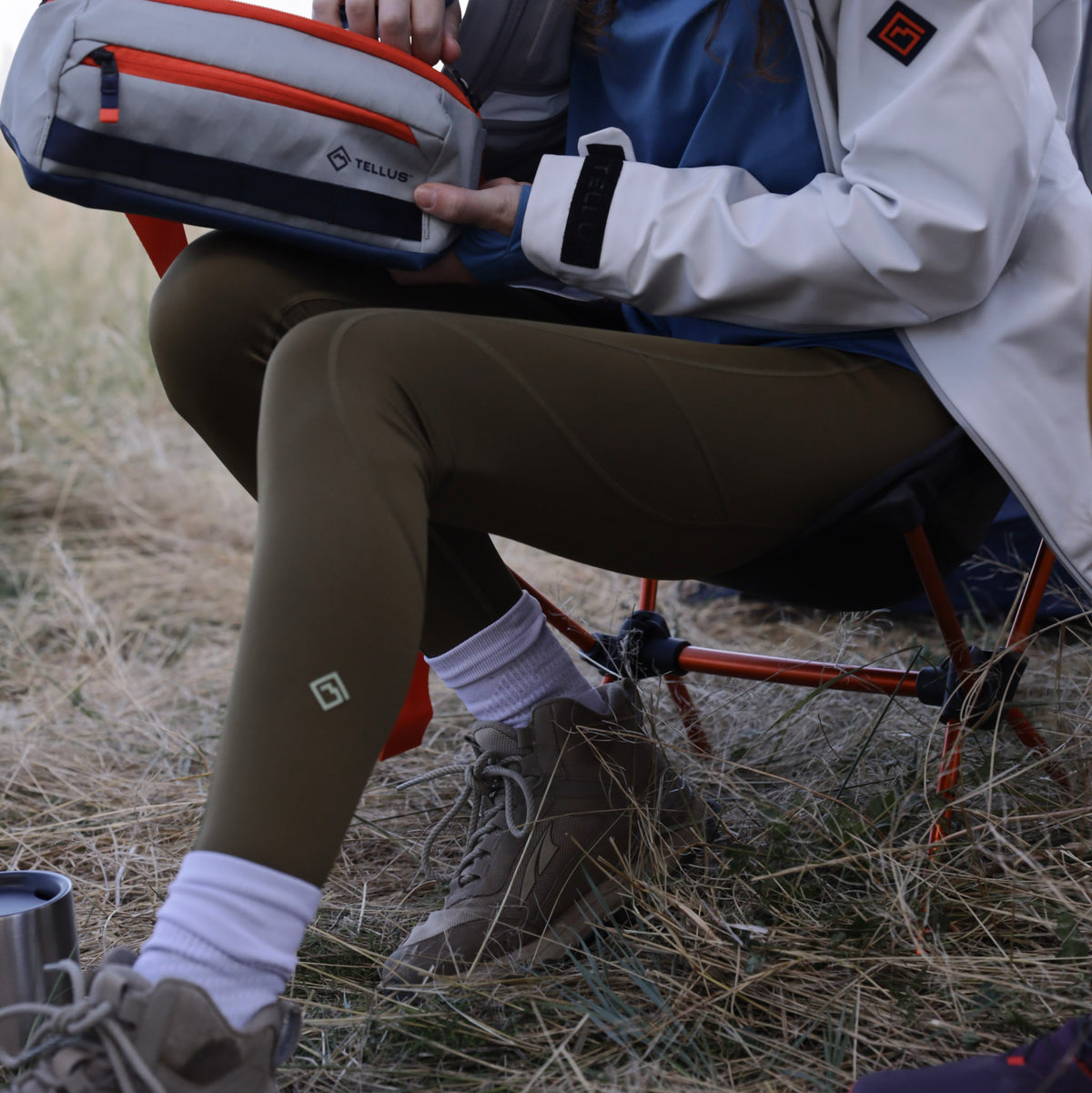 A woman sitting outdoors on a camp chair holds a gray and orange Neota Sling Bag. She is wearing olive green Sunset Ridge Performance leggings, white socks and brown hiking boots.