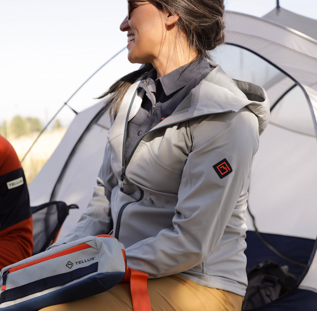 A woman wearing a light gray Timberline Soft Shell hooded jacket sits beside a white and navy tent, holding a gray, navy, and orange Tellus Neota sling bag.