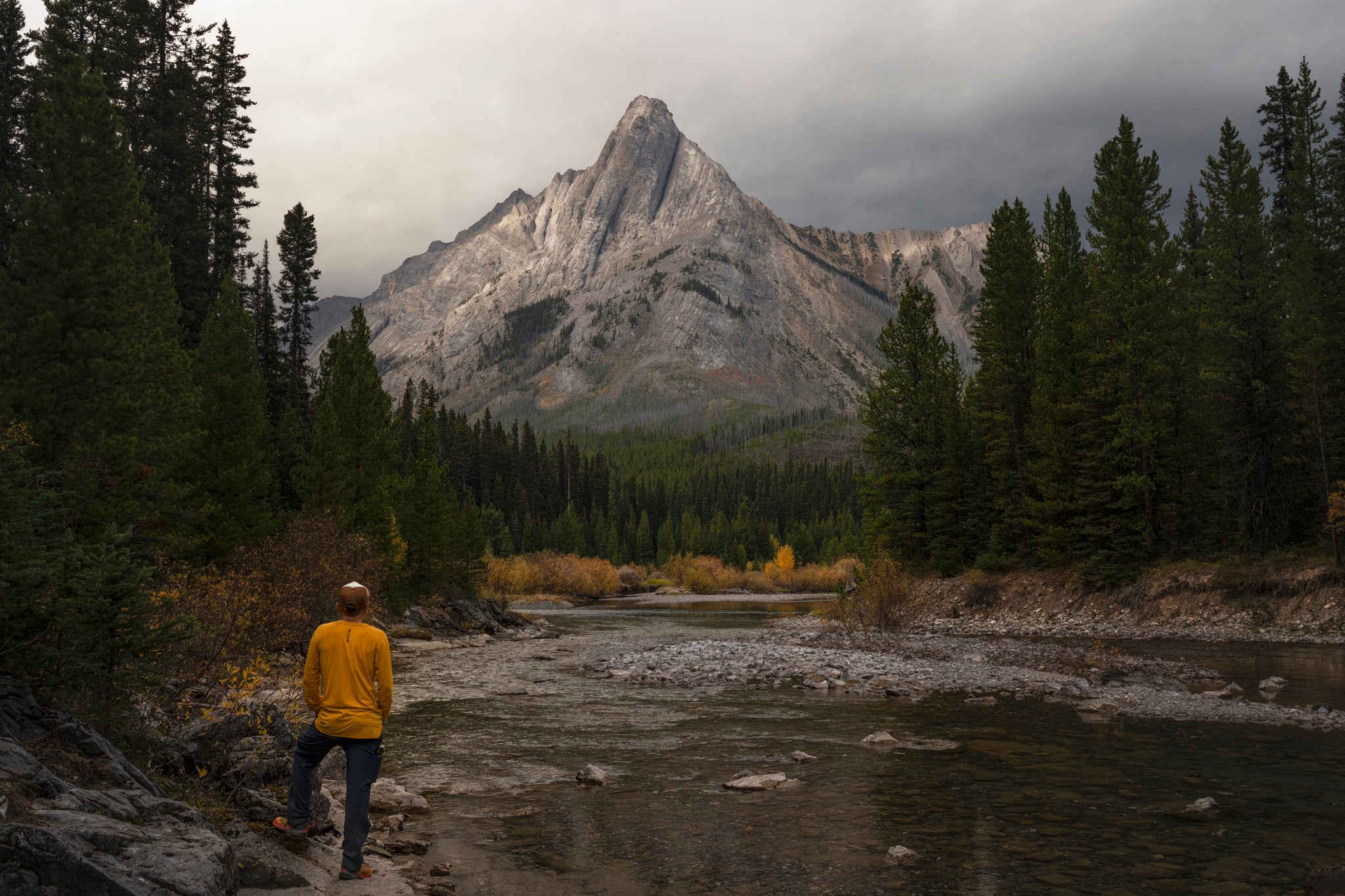 Person in a yellow jacket standing near a river with a mountain in the background