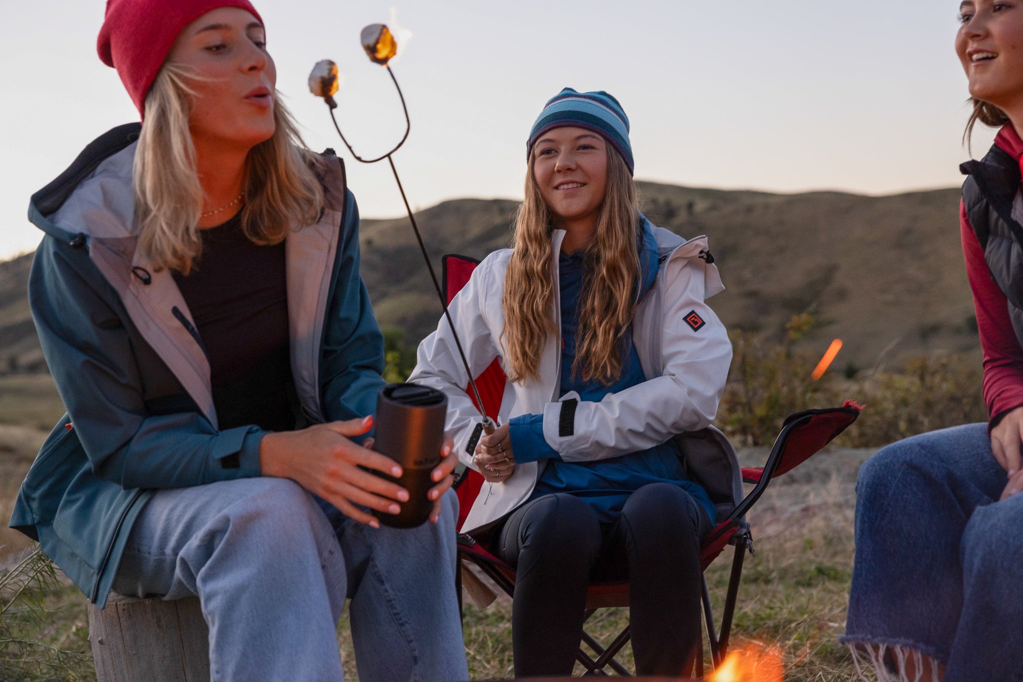 Three people sitting outdoors in a mountainous area, roasting marshmallows and enjoying a campfire.