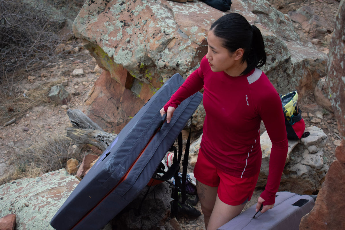 A woman in a red Poudre Trail Performance long-sleeve athletic shirt and matching Sunset Ridge Performance shorts carries climbing gear, including a large gray pads through a rocky outdoor terrain.