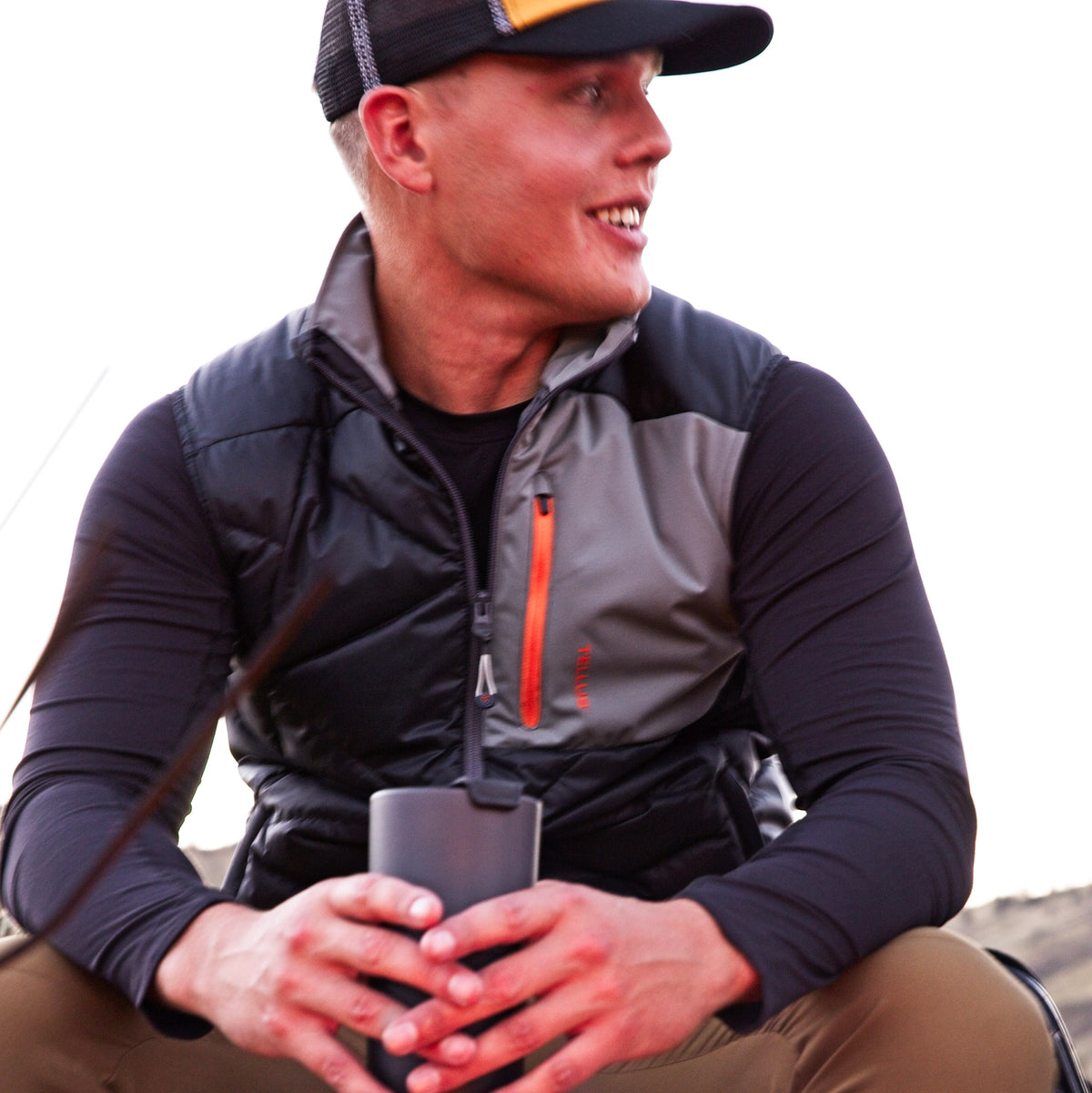 A man wearing a black and gray Byers Peak Recycled Down vest over a black Poudre Trail Performance long-sleeve shirt, holding a black insulated tumbler. He is wearing a black and yellow Tellus trucker cap, set against a bright, overexposed outdoor background.