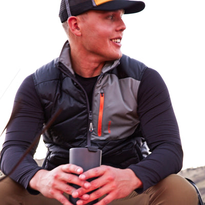 A man wearing a black and gray Byers Peak Recycled Down vest over a black Poudre Trail Performance long-sleeve shirt, holding a black insulated tumbler. He is wearing a black and yellow Tellus trucker cap, set against a bright, overexposed outdoor background.