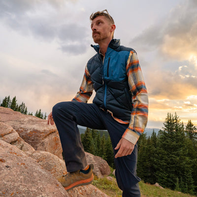 A man wearing a blue and navy quilted Byers Peak Recycled Down vest over an orange and beige plaid Foothills Flannel shirt, Blue Trail Ridge Trek hiking pants, and hiking boots, posing on a rocky mountain peak with a forest and sunset in the background.