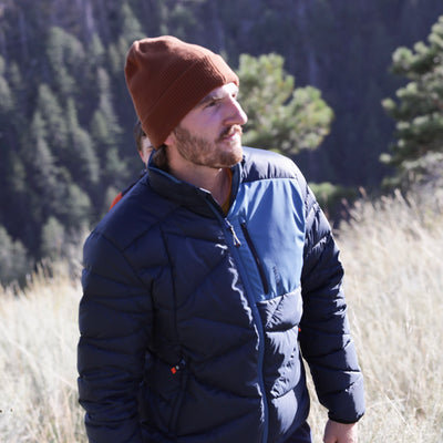 A man wearing a brown knit Tellus beanie and a navy blue Byers Peak Jacket with a lighter blue yoke, standing outdoors in a grassy field with trees in the background.