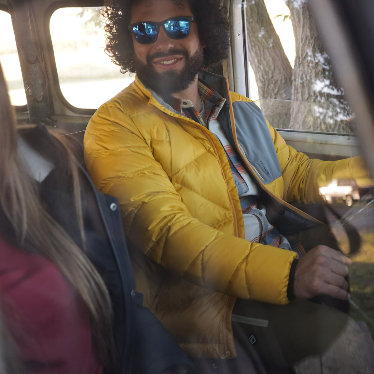 A man with a beard and sunglasses wearing a yellow Byers Peak Recycled Down jacket with gray panels, sitting inside a vehicle.