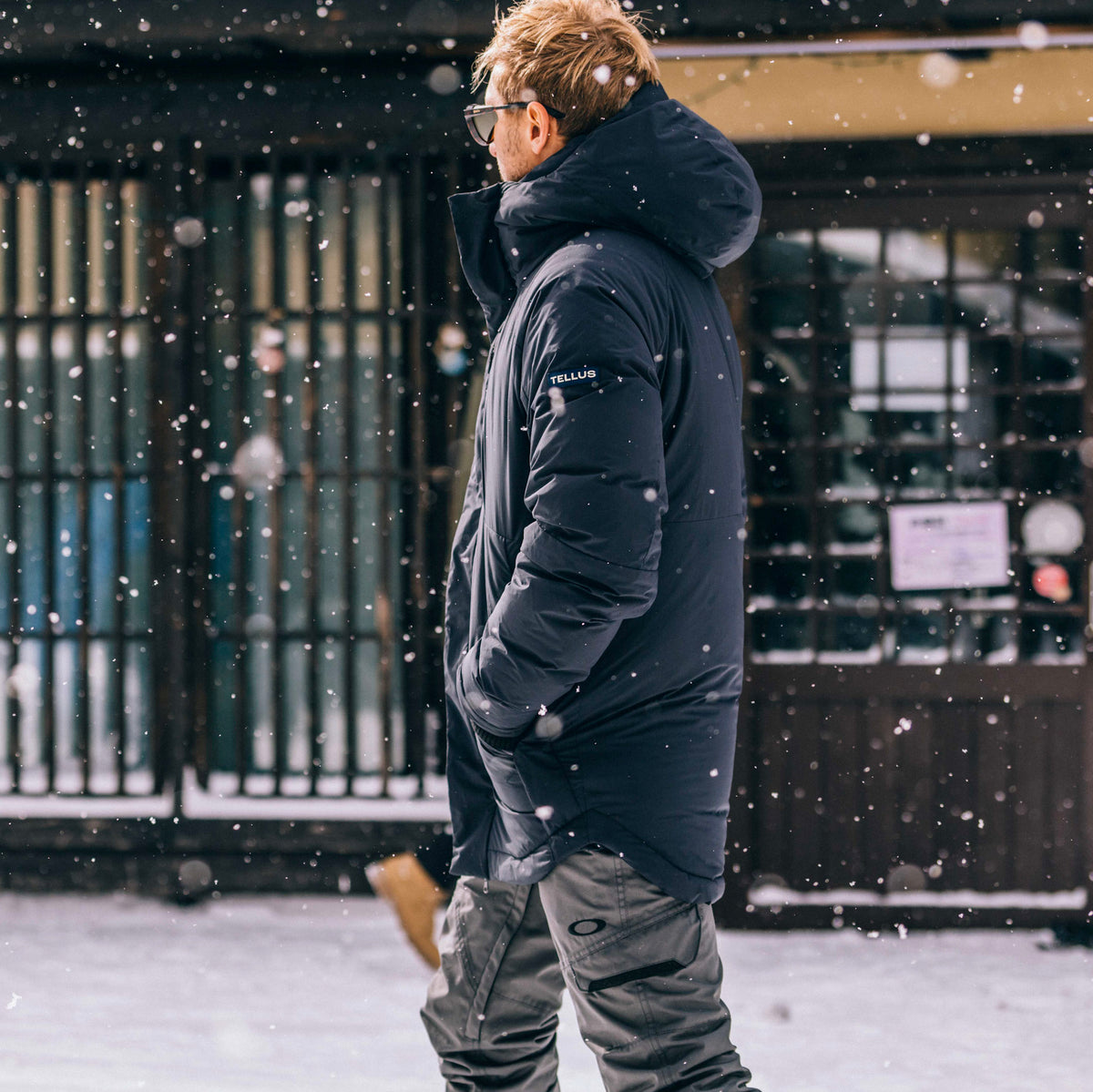 A man wearing a black hooded Cameron Pass Parka jacket and gray pants walking outdoors in the snow in front of a building.