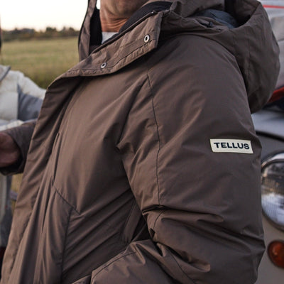 A close-up of a man wearing a brown hooded Cameron Pass Parka jacket outdoors. A small white rectangular patch on the upper right sleeve reads TELLUS, which is the visible brand.