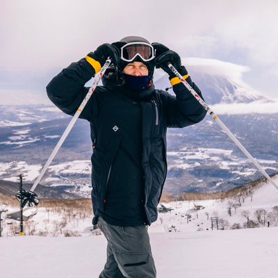A skier wearing a black Cameron Pass Parka jacket, gray pants, black gloves, and goggles stands on a snowy mountain slope with ski poles, looking toward the camera. A snow-covered mountain and cloudy sky are visible in the background.