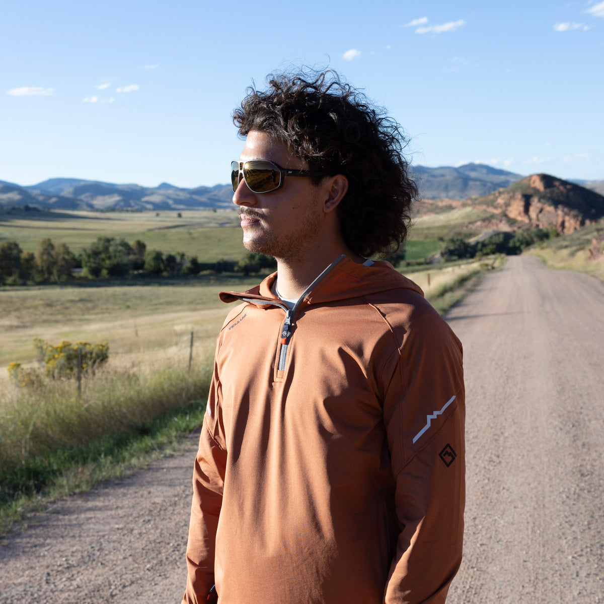 A man with curly dark hair and sunglasses stands on a dirt road wearing an orange Horsetooth Hoodie Quarter Zip set against a backdrop of rolling green hills and mountains under a blue sky.