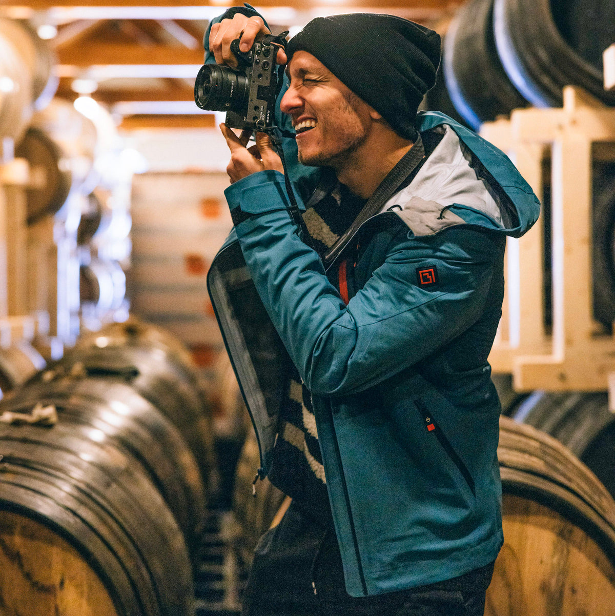 A man wearing a blue Long's Peak Waterproof Rain Shell jacket, black beanie, and striped sweater holds a camera in a wine cellar with wooden barrels in the background.