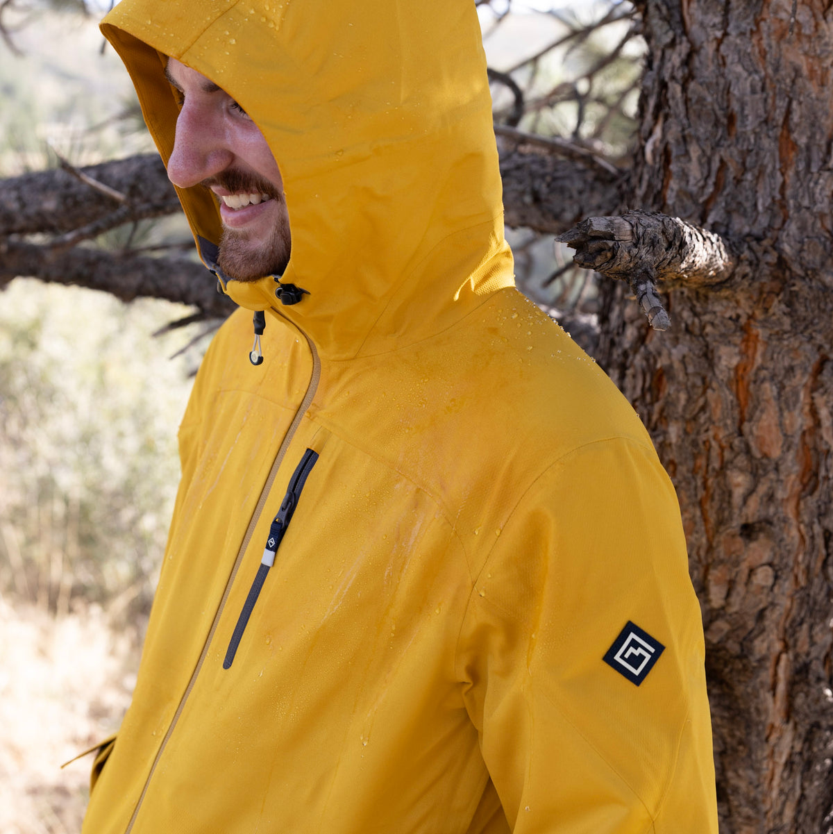 A man wearing a bright yellow Long's Peak Waterproof Rain Shell hooded rain jacket stands outdoors next to a tree, with water droplets visible on the jacket.