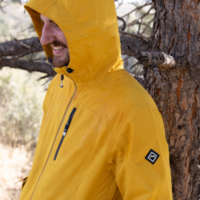 A man wearing a bright yellow Long's Peak Waterproof Rain Shell hooded rain jacket stands outdoors next to a tree, with water droplets visible on the jacket.