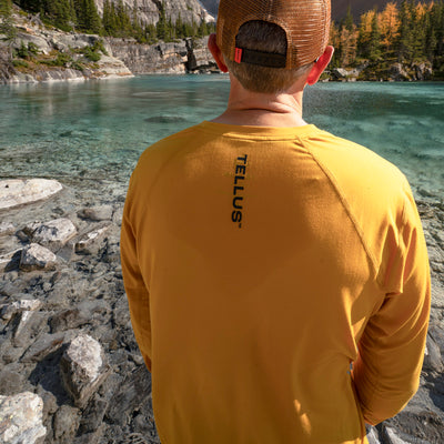 Back of man wearing yellow Tellus Poudre Trail Performance long sleeve shirt looking over a lake with mountains and trees in background.