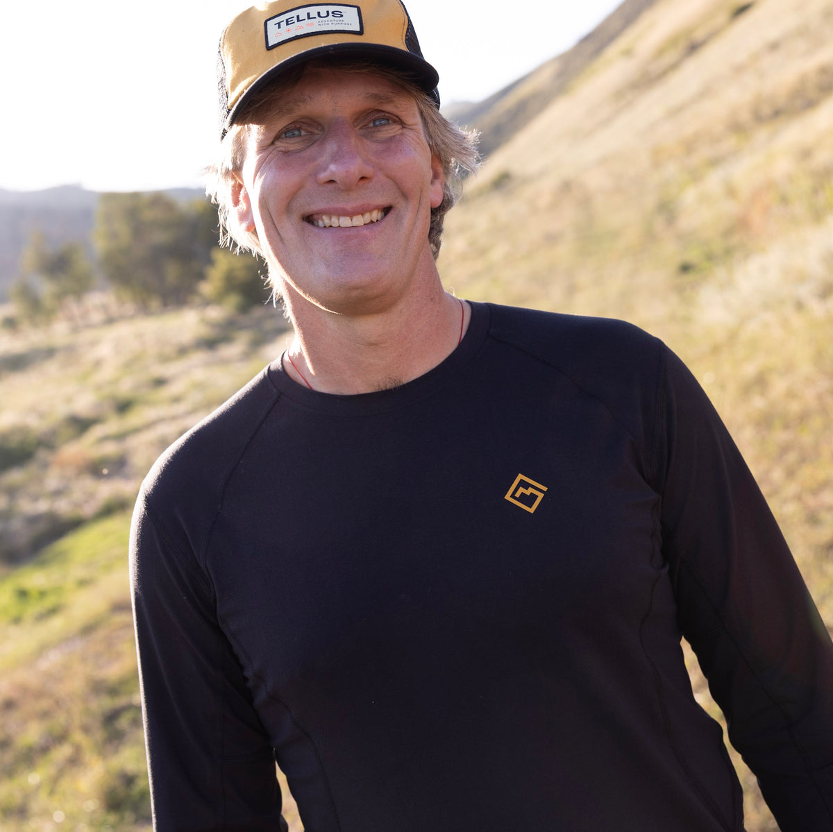 A smiling man outdoors on a grassy hill wearing a black long-sleeve Poudre Trail Performance long sleeve athletic shirt with a small gold logo on the chest and a tan and black Tellus trucker hat.