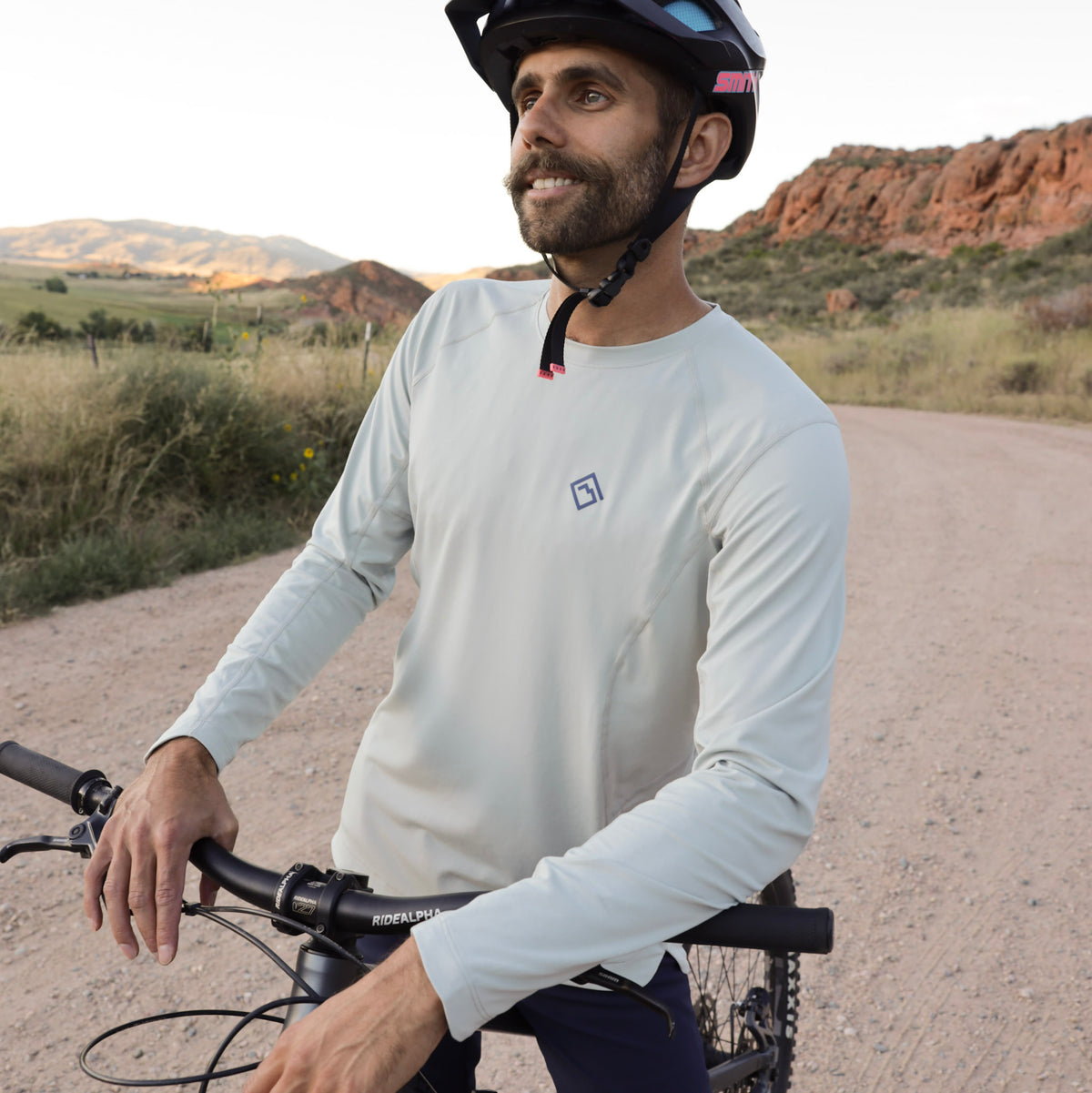 Man on bicycle on dirt road wearing white Tellus Poudre Trail Performance long sleeve shirt with mountains in the background.