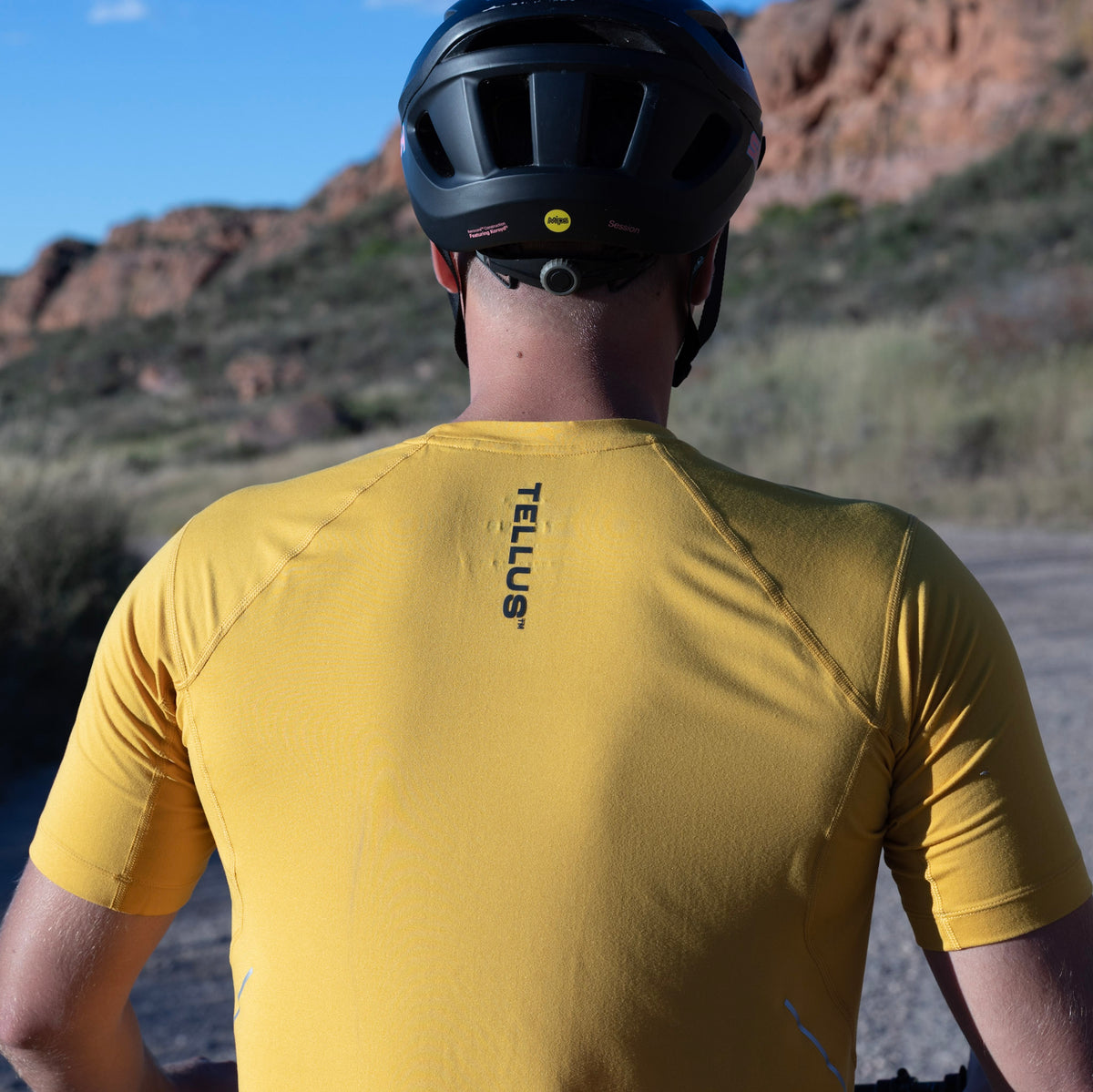 A man viewed from behind wearing a yellow Poudre Trail Performance short sleeve shirt with 'TELLUS' printed vertically on the back, along with a black helmet, set against a desert canyon background.