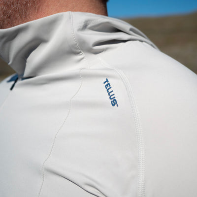 Close-up of a person wearing a light gray Satanka Cove Hoodie sun shirt outdoors, showing the shoulder and neck area with a blue TELLUS logo on the upper chest.