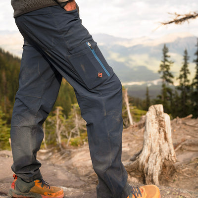 A man outdoors wearing dark navy Trail Ridge Trek Pant with a blue zippered pocket and orange hiking boots, standing on a rocky mountain trail with trees and a forest valley in the background.