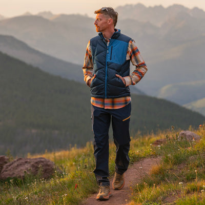 A man hiking on a mountain trail wearing a navy blue quilted Byers Beak Recycled Down vest over a plaid Foothills Flannel shirt and navy Trail Ridge Trek hiking pants, with layered mountain ranges in the background.