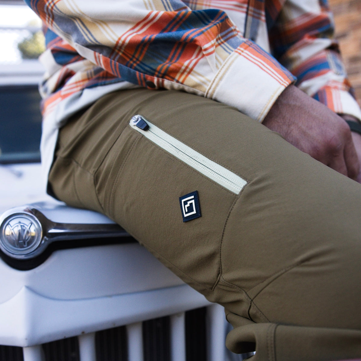 Close-up of a man wearing olive brown Trail Ridge Trek pants with a diagonal zippered pocket and a small square Tellus logo patch, seated on the front of a white vehicle.