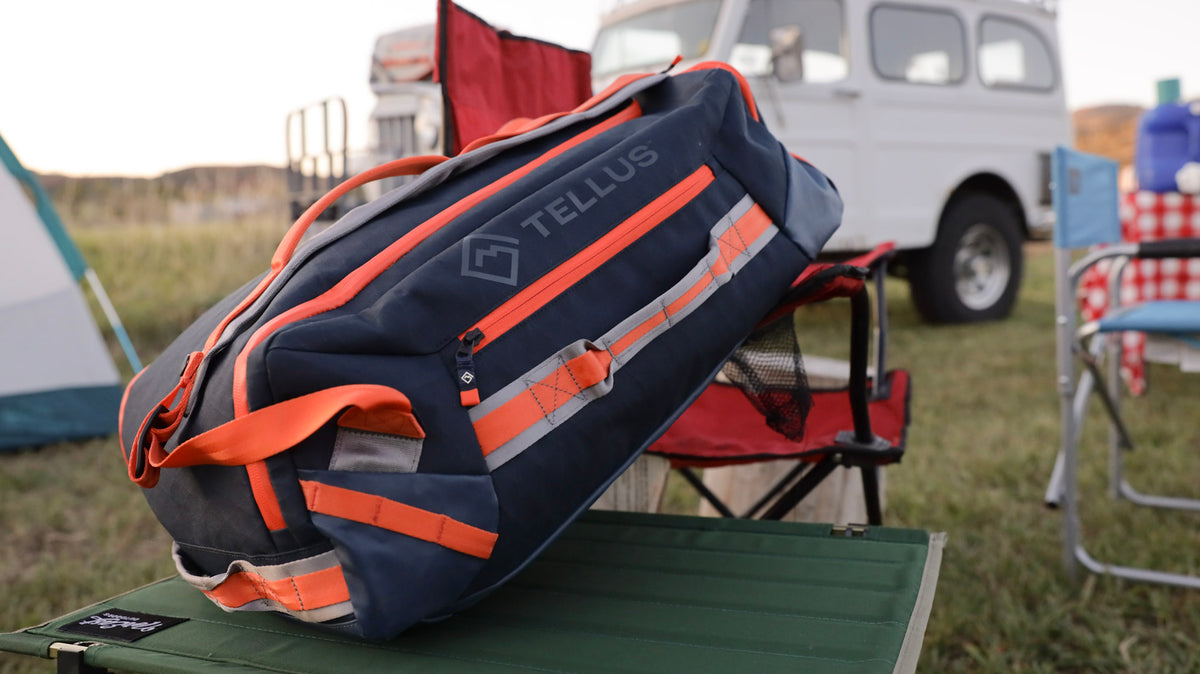 Tellus Cache Carryall duffle leaning on a table and chair in a campsite with a classic SUV in the background.