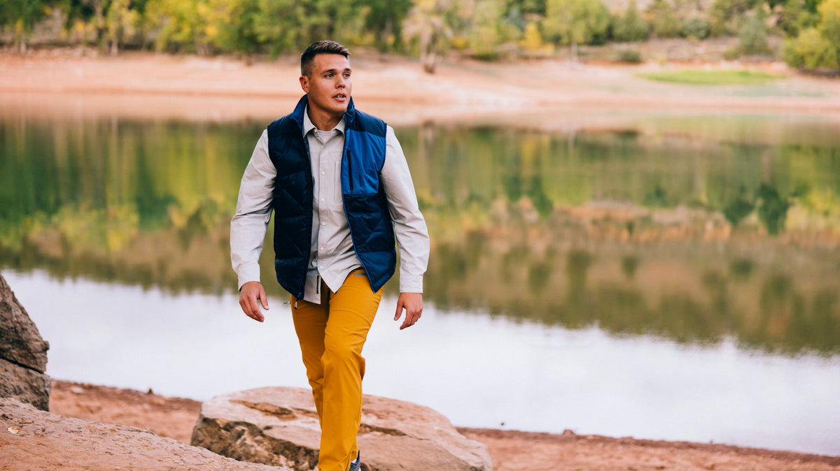 Man wearing dark blue Byers Peak Recycled Down vest, light gray Spring Creek Button Down and mustard Trail Ridge Trek pants with a lake and trees behind him.