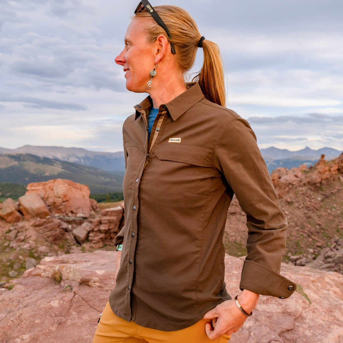 A woman wearing a brown Foothills Flannel button-up shirt and tan pants stands outdoors on a rocky mountain landscape, looking to the side.
