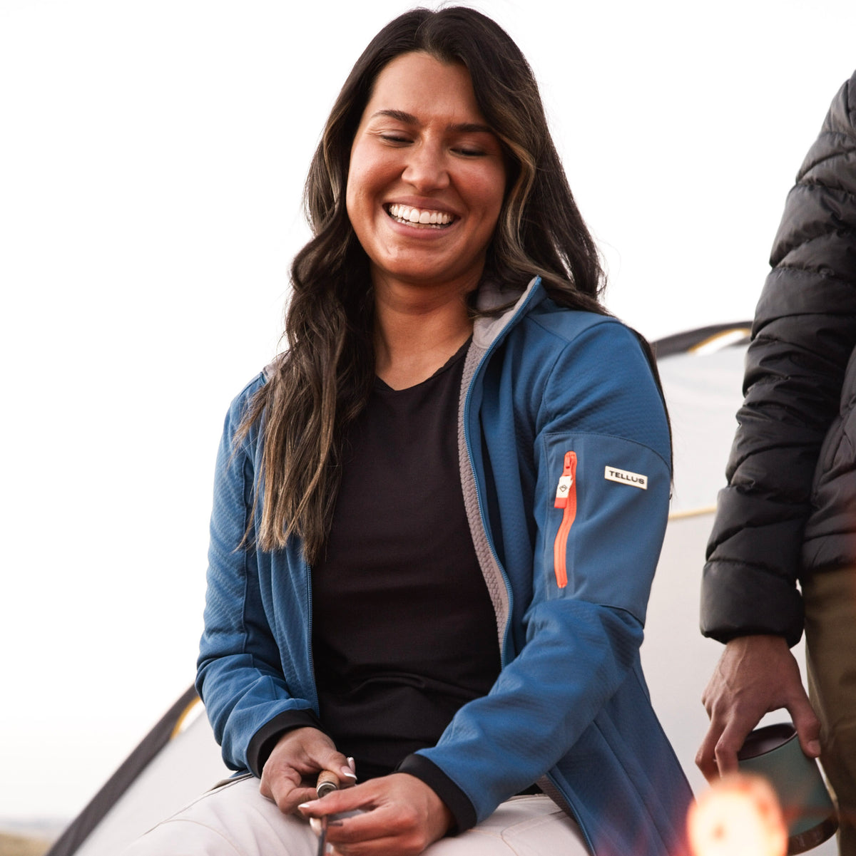 A woman wearing a blue Front Range Fleece zippered jacket with a black shirt, smiling outdoors next to a campfire.
