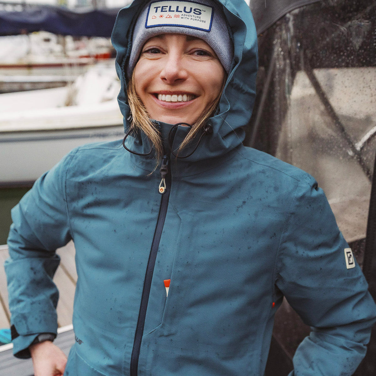 A woman smiling while wearing a blue Tellus Long's Peak waterproof rain shell jacket and a gray beanie with a Tellus logo patch, standing outdoors in the rain near a boat.