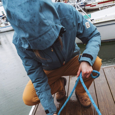 A woman is crouching on a wooden dock by the water, wearing a blue hooded Long's Peak Waterproof Rain Shell jacket while holding a blue rope. Boats and water are visible in the background.