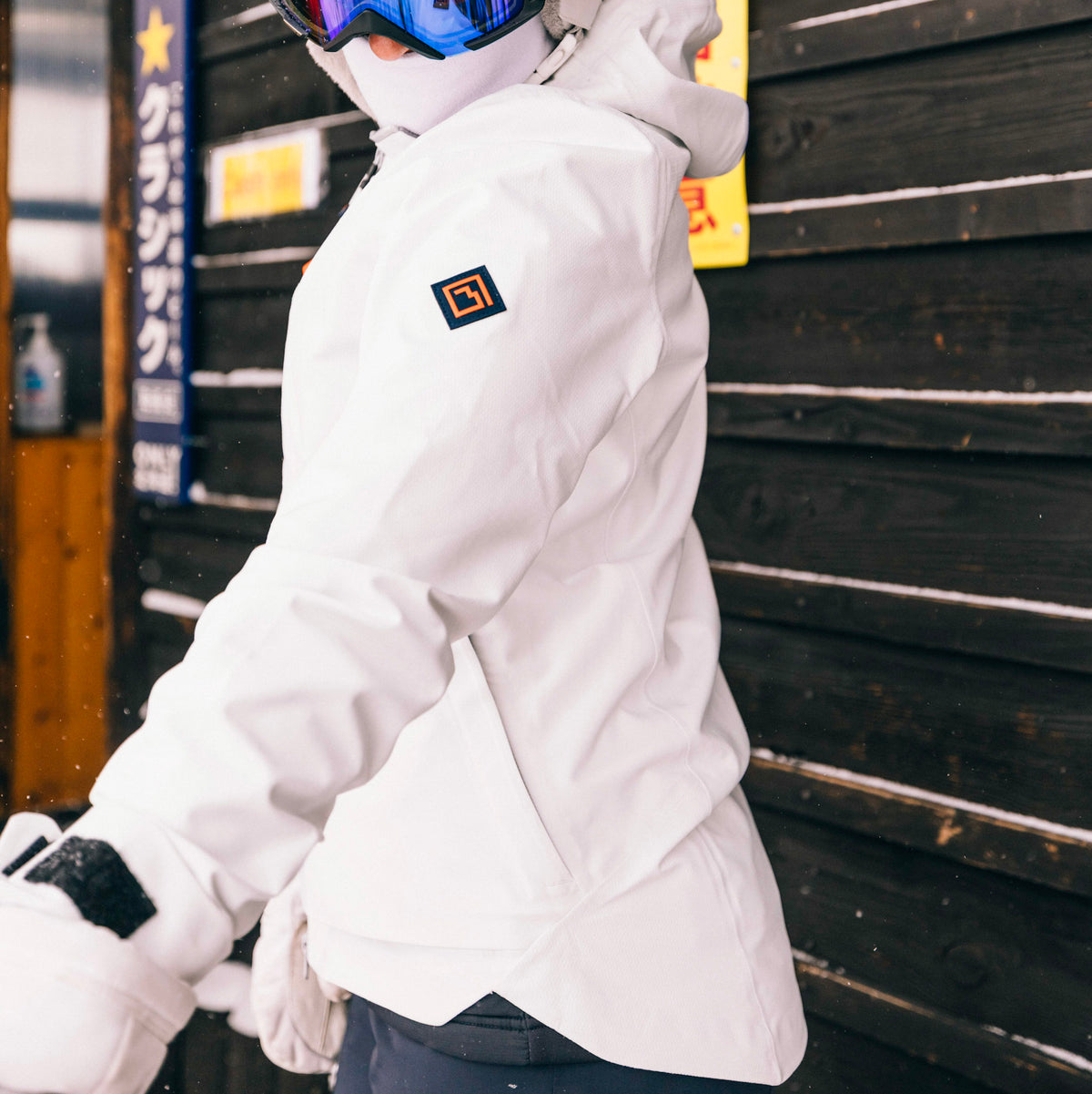 A woman wearing a white Long's Peak Waterproof Rain Shell jacket, white gloves, and blue ski goggles stands in front of a dark wooden wall. The jacket has a small black and orange Tellus logo patch on the left shoulder. 