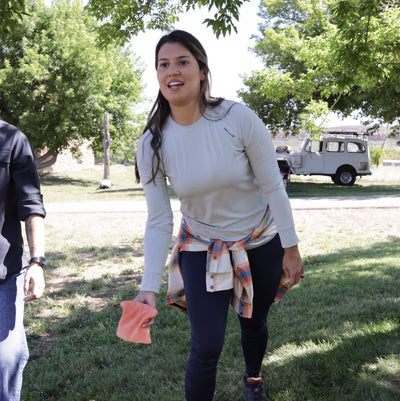 A woman outdoors on a grassy area with trees, wearing a gray Poudre Trail Performance long sleeve shirt and dark blue Sunset Ridge Performance leggings, playing cornhole. 
