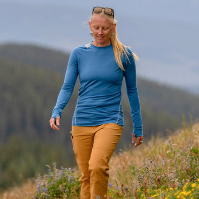 A woman hiking outdoors in a blue Poudre Trail Performance long-sleeve shirt and tan Trail Ridge Trek hiking pants, with blurred hills in the background.