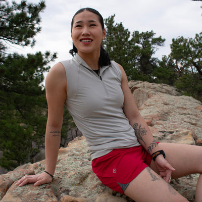 Woman wearing gray Satanka Cove Tank sitting and jester red Sunset Ridge Performance Shorts on rock with pine trees in background