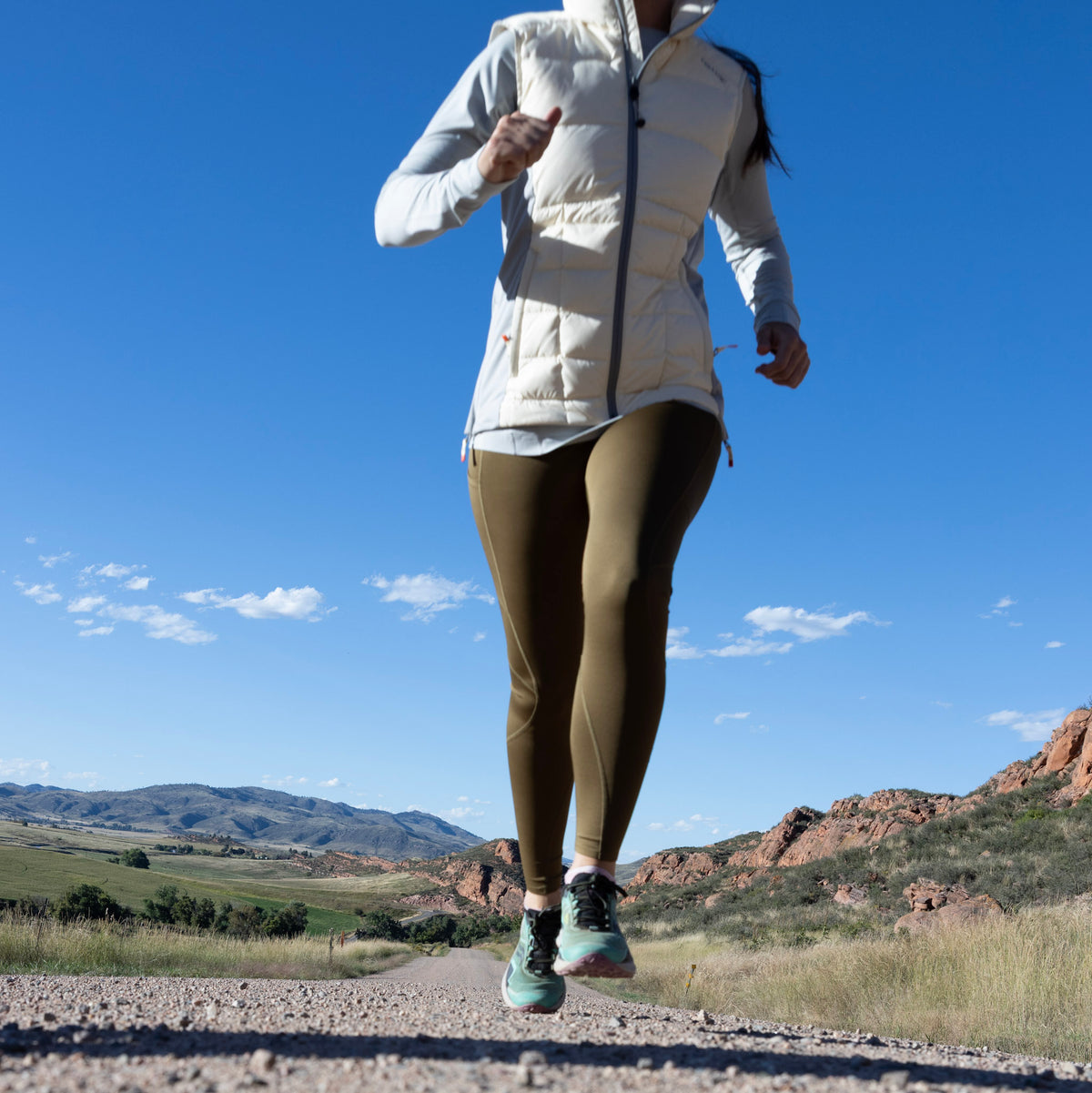A woman running along on a dirt road wearing a white Byers Peak Recycled down vest over a light gray long-sleeve top, olive green Sunset Ridge Performance leggings, and mint green running shoes, with a blue sky and rocky hills in the background.