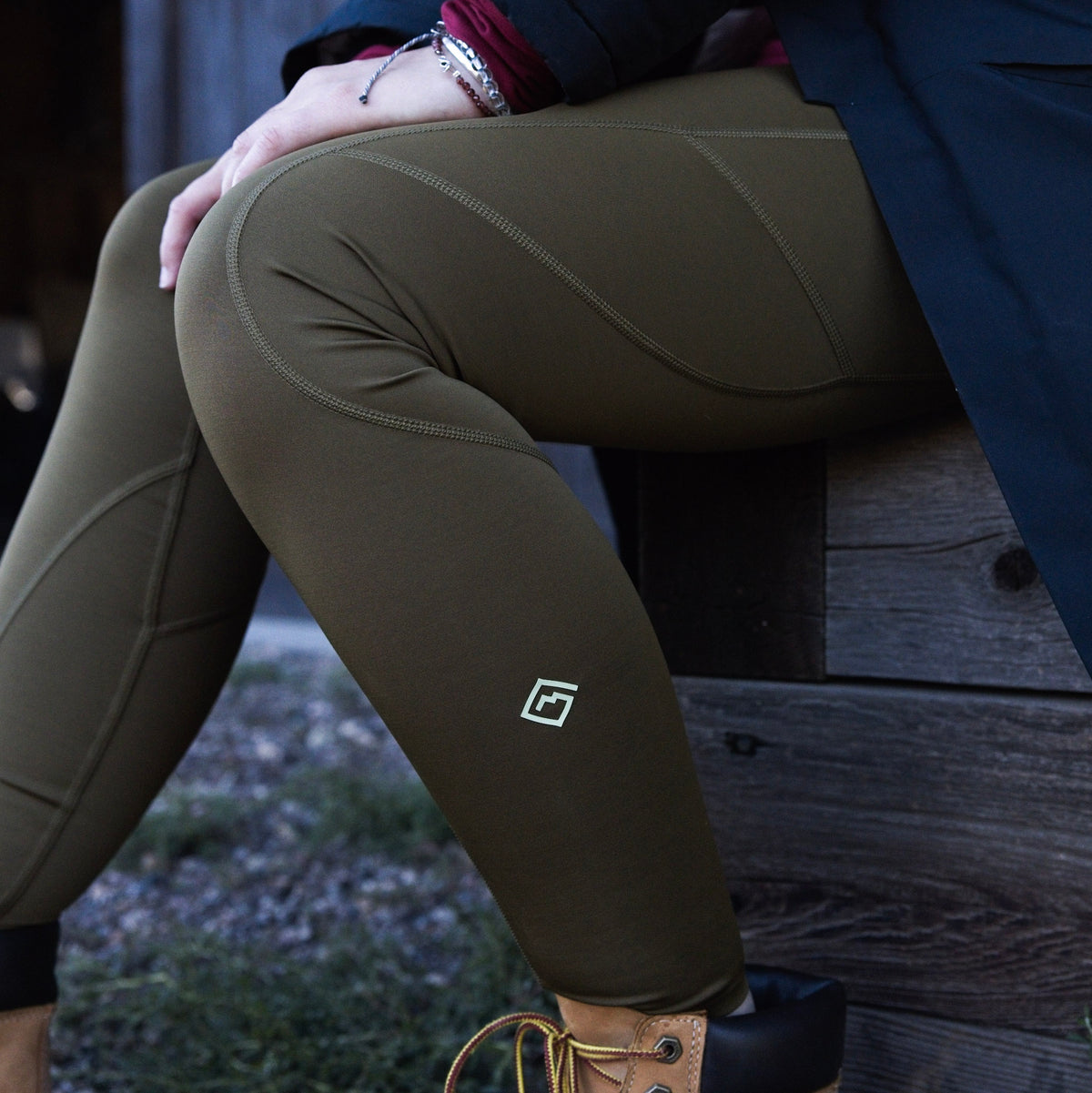 A close-up lifestyle shot of a woman wearing olive green Sunset Ridge Performance leggings with a small white Tellus logo on the lower left leg, sitting on a rustic wooden bench outdoors.