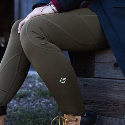 A close-up lifestyle shot of a woman wearing olive green Sunset Ridge Performance leggings with a small white Tellus logo on the lower left leg, sitting on a rustic wooden bench outdoors.
