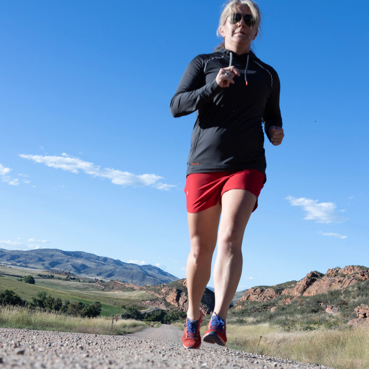 Woman running in desert with hills in background wearing red Sunset Ridge Performance shorts and dark gray Horsetooth Performance Hoodie.