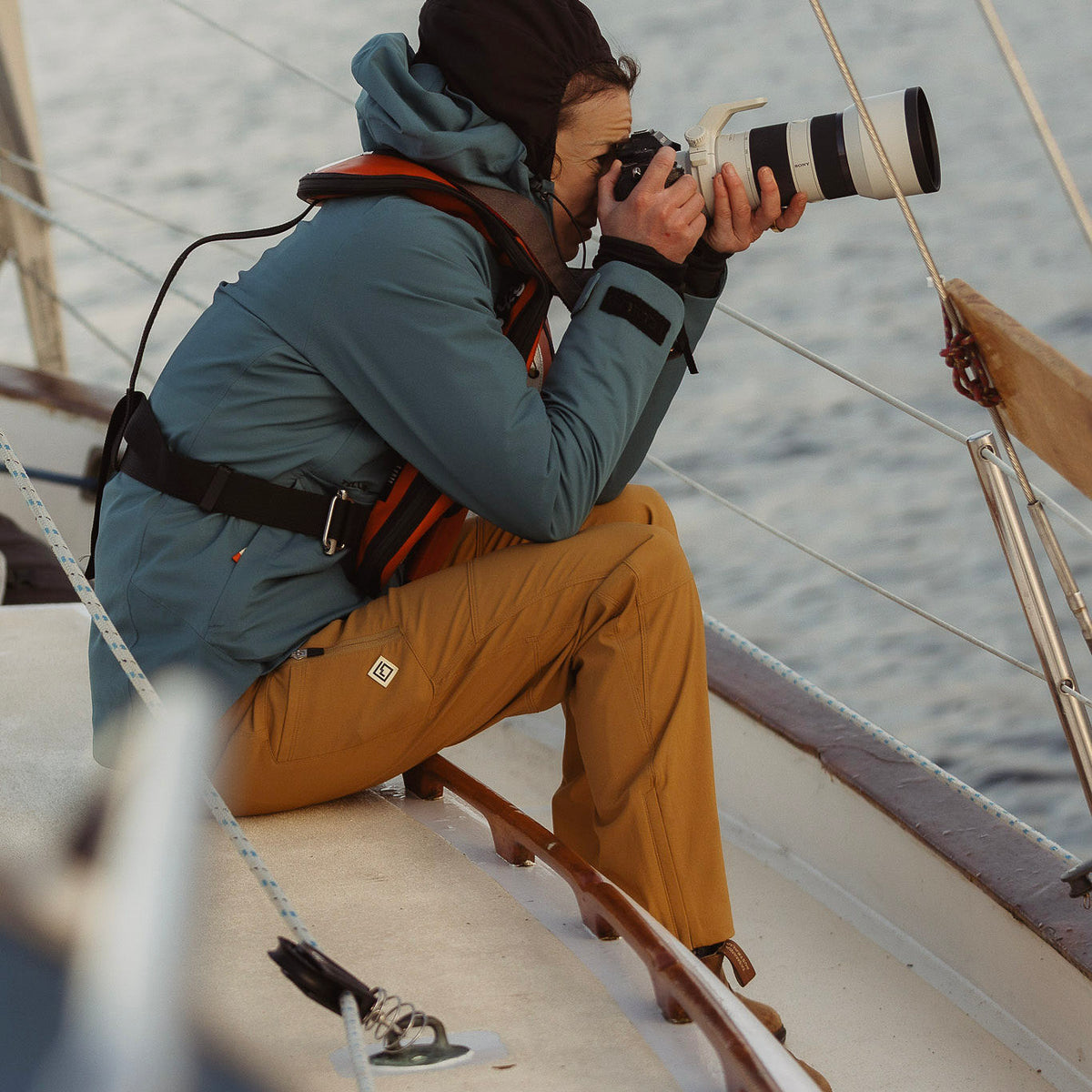 A woman crouching on a sailboat deck holding a camera with a long telephoto lens, wearing tan Trail Ridge Trek pants.