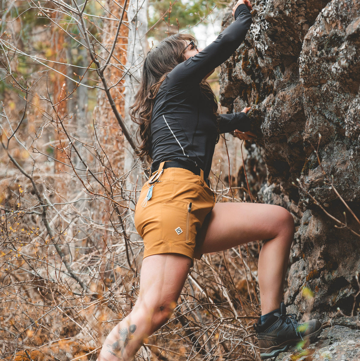 A woman climbing a rock face in a forest, wearing a black Poudre Trail Peformance long-sleeve top and tan Trail Ridge Trek shorts with a small white Tellus logo patch.