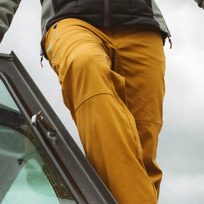 A close-up of a woman wearing mustard yellow Trail Ridge Trek pants stepping onto a boat against an overcast sky.